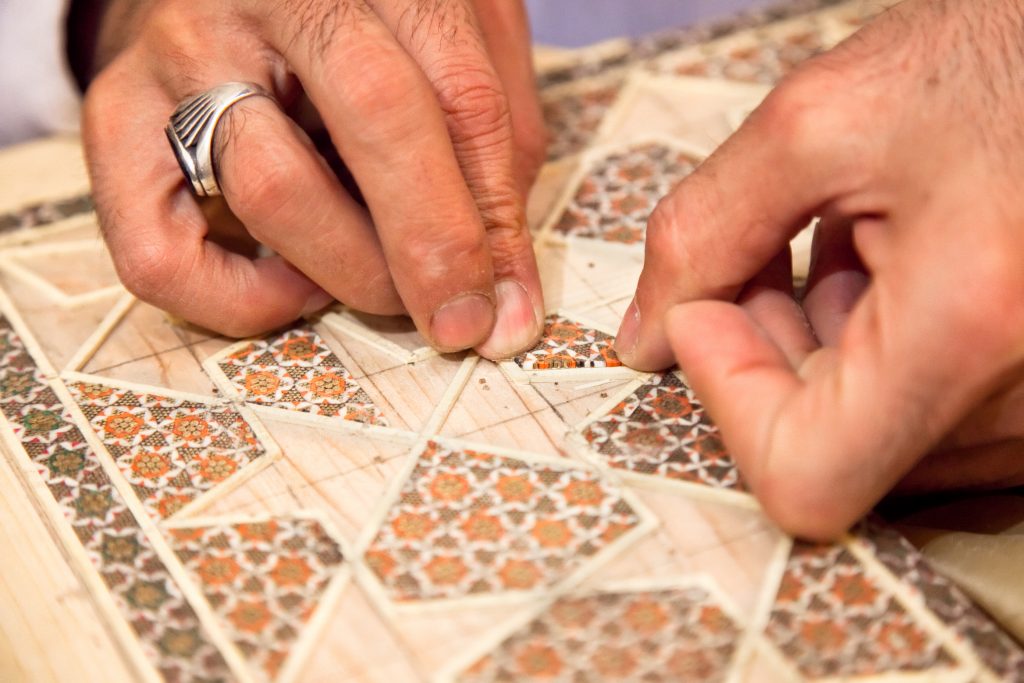 A Persian craftsman works meticulously with his hands, carefully arranging wooden pieces to create an intricate design on a piece of art using the traditional marquetry technique. His steady and skilled fingers create patterns that demonstrate his mastery of the craft, showcasing the beauty and elegance of Persian artistry.
