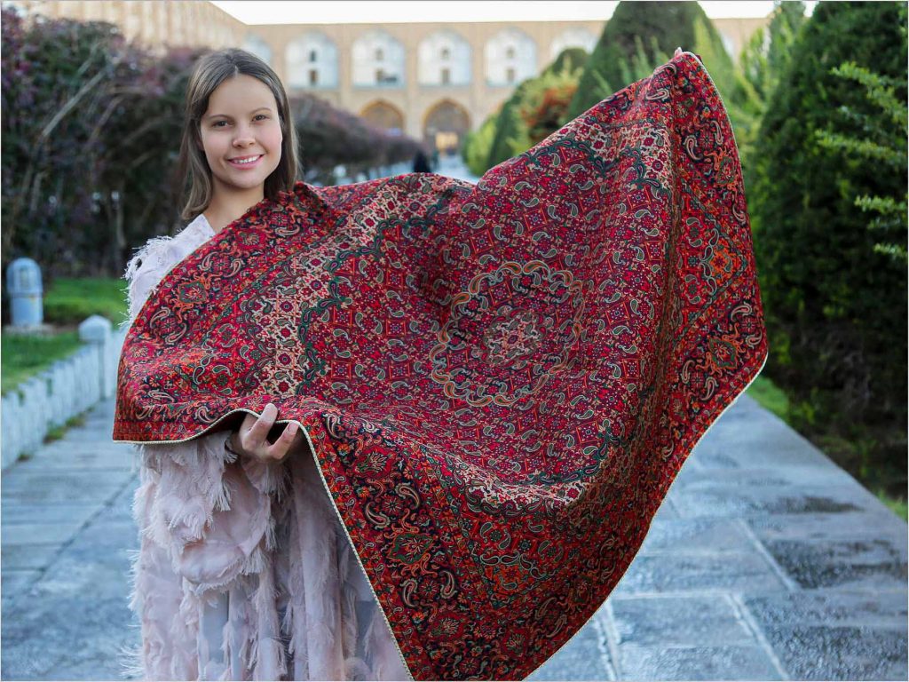 A woman holding an authentic Persian textile Termeh tablecloth with intricate red and gold patterns.