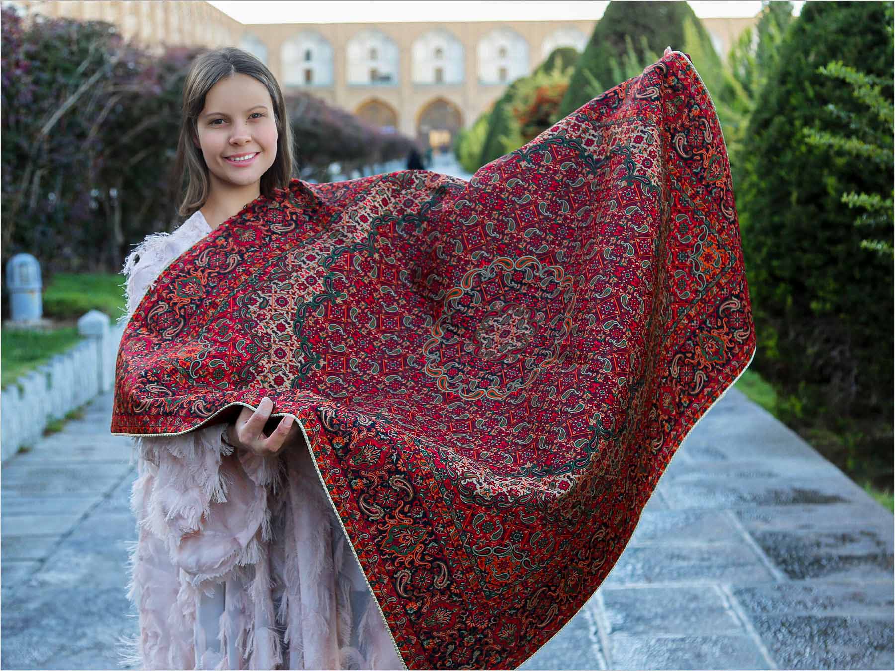 A woman holding an authentic Persian textile Termeh tablecloth with intricate red and gold patterns.