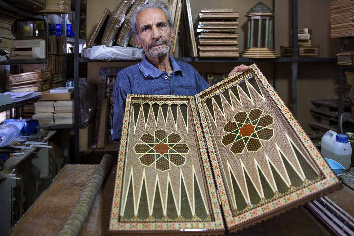 A proud Persian craftsman holds his masterpiece, a beautifully crafted wooden backgammon board. Using the intricate marquetry technique, the wooden pieces are inlaid with intricate designs and patterns, showcasing the craftsman's attention to detail and skill in his trade.