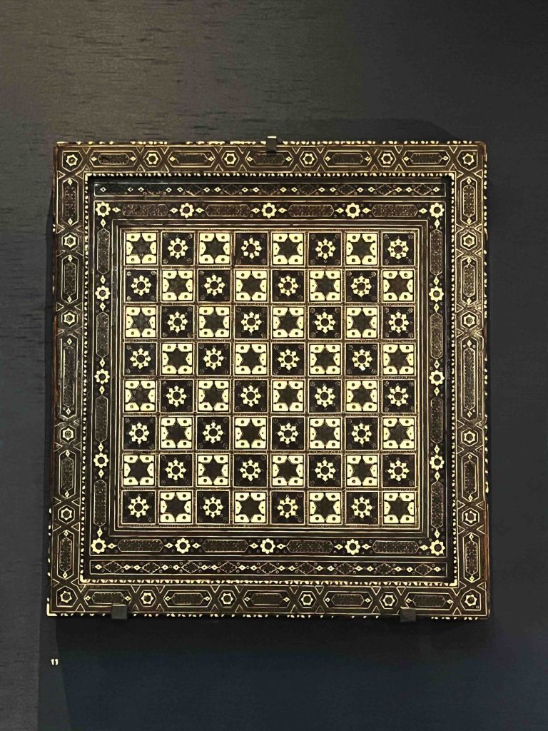 A photograph of an antique chess board from Northwest India on display at the British Museum. The board is crafted from wood and inlaid with bone or ivory and green paste in an intricate design dating back to the 17th or 18th century. The craftsmanship and materials reflect the luxury and cultural heritage of the Indian subcontinent. History of Board Games