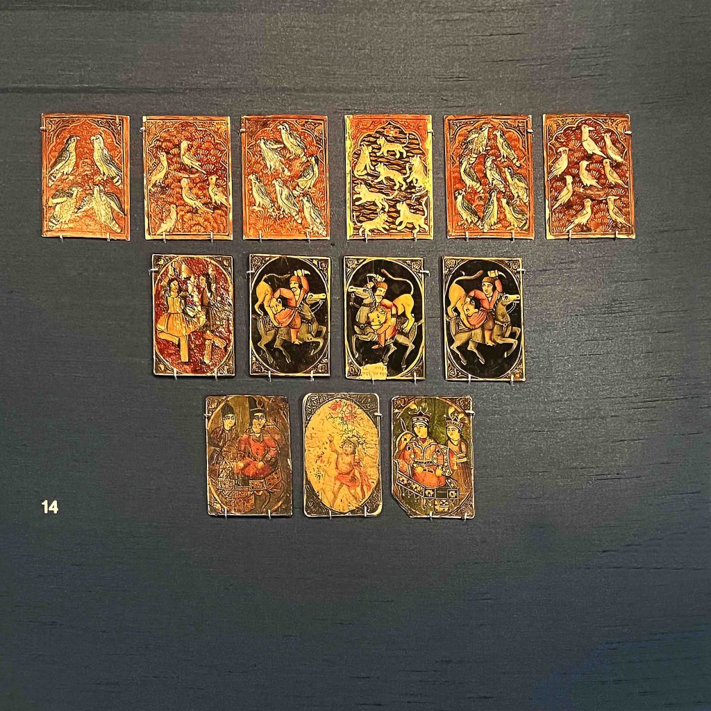 A photograph of Iranian playing cards from the 1800-1900s on display at the British Museum. The cards feature intricate designs depicting Persian epics, everyday life scenes, foreigners, and erotic scenes, with different animals or birds used to distinguish the suits. These cards offer insight into the gaming and artistic traditions of Iran during this time period. History of Board Games