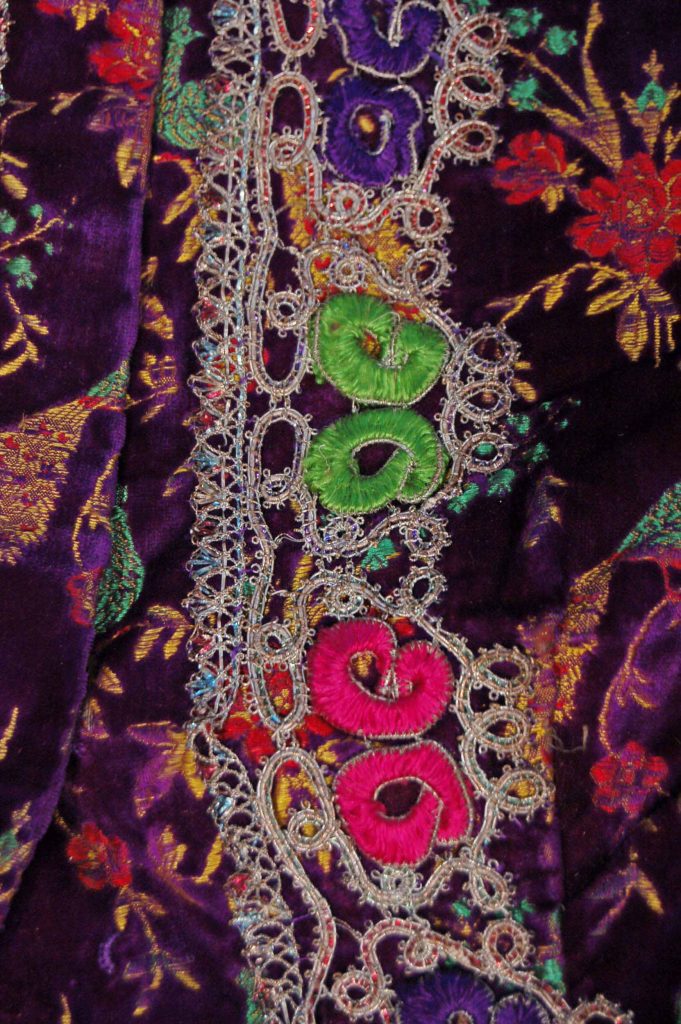 This exquisite purple velvet jacket features intricate woven designs of peacocks and colorful flower sprays, showcasing the vibrant artistry of Persian textiles. The jacket is further enhanced with embroidered and couched designs along the front edges, hem, collar, and cuffs, creating a striking visual appeal. Acquired in Iran, this jacket represents the rich cultural heritage and skilled craftsmanship characteristic of traditional Iranian attire.