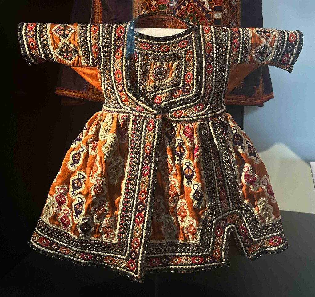 A photo of a child's vibrant orange silk outfit in Baluchistan, intricately embroidered with silk thread stitches, ricrac, sequins, and mirror work (shisho). The lavish embellishments, including glittering mirrors and sequins, serve a dual purpose of enhancing the garment's allure and safeguarding the wearer from misfortune and envy.