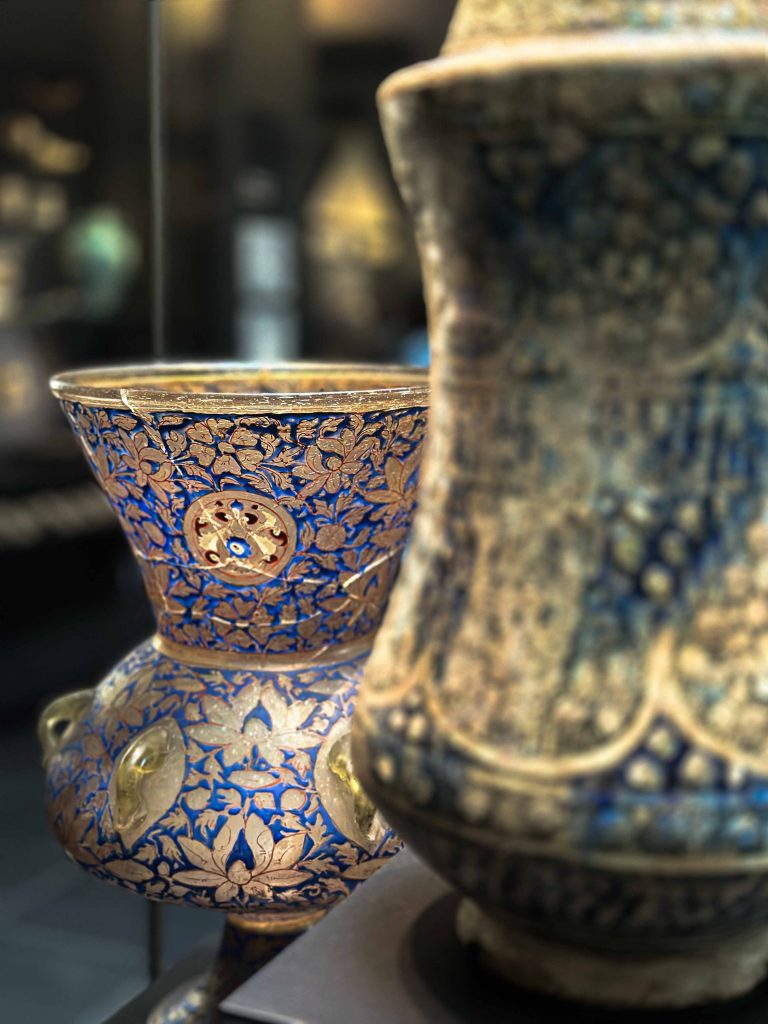 Image of a mid-14th-century Mamluk mosque lamp with a dark blue enamelled background. The lamp features meticulously crafted circular medallions, each bearing lotus motifs that speak to a rich tapestry of cultural exchange and artistic evolution in the Islamic world.