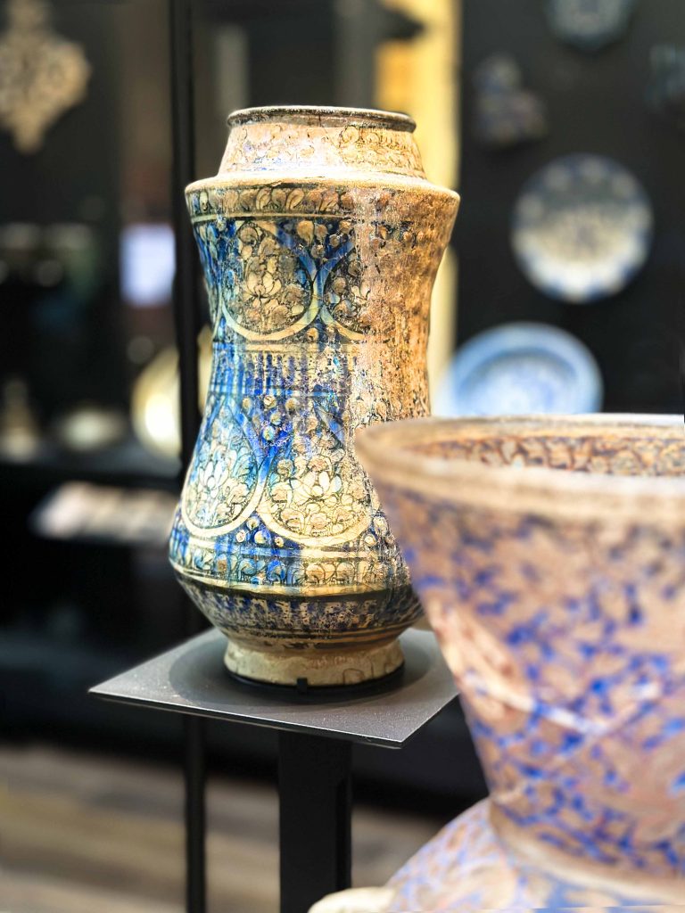 Image of a stonepaste albarello from the Ilkhanid Sultanabad group dating 1300-1400, showcasing a vibrant blue glaze and intricate slip-painted lotus motifs in grey-green, encapsulating the early stages of Persian enamelling or 'Meenakari'