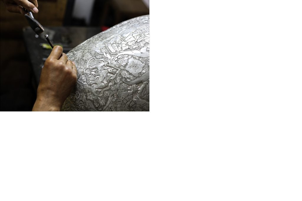 Persian craftsman working on a decorative vase using Ghalam Zani technique, holding a mallet and chisels.