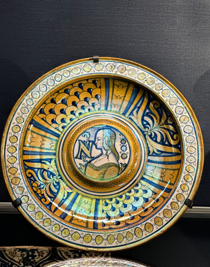 A Renaissance dish from Deruta features a blue-painted female portrait with the letter "N," encircled by intricate patterns of scales and stylized foliage, all accentuated with a brownish lustre—an embodiment of Islamic influence on lustre ceramics and a significant piece in the history of enamelling.