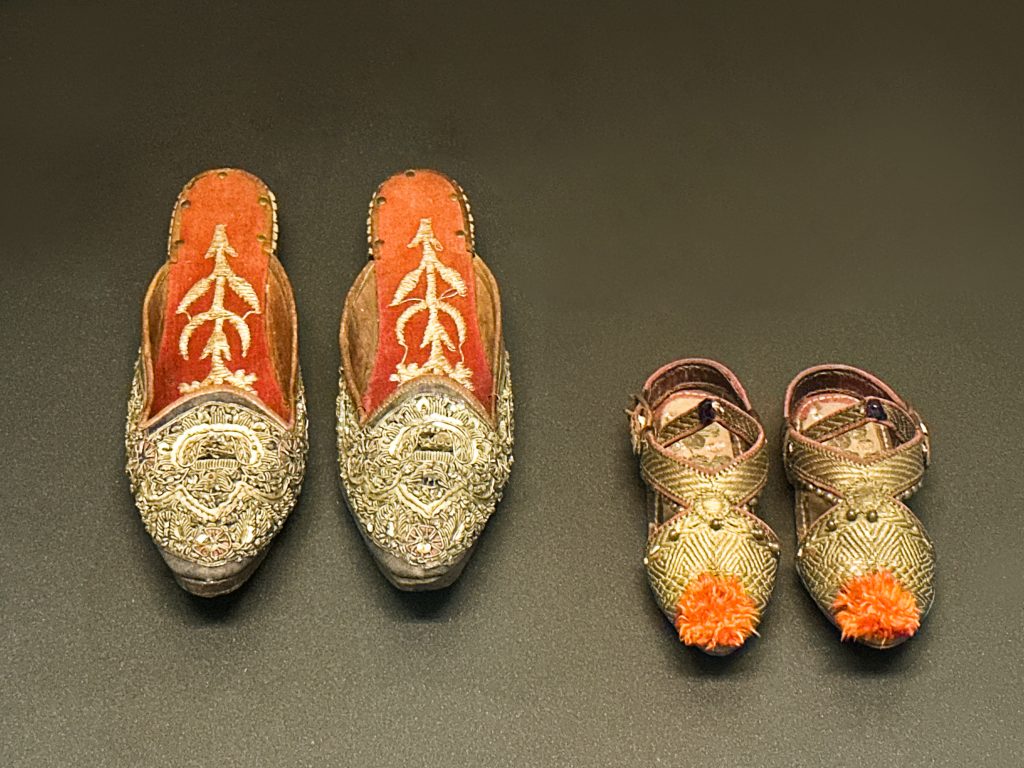 A pair of blue velvet lady's slippers embroidered with gold-thread, and a pair of children's leather sandals adorned with gilt embroidery, glass beads, and silk pom-poms, both exemplifying Ottoman Empire Attire.