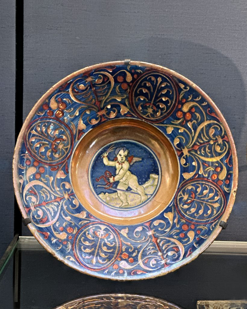 A golden-lustred Renaissance bowl from Gubbio, showcasing a winged cherub riding a hobby horse and adorned with intricate blue and red patterns on the rim and reverse, reflects the Islamic influence on lustre ceramics and contributes to the history of enamelling.