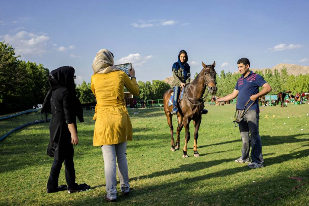 Young Iranian girl on a horse with an assisting man, while two other girls photograph the scene in an Iranian horse farm.