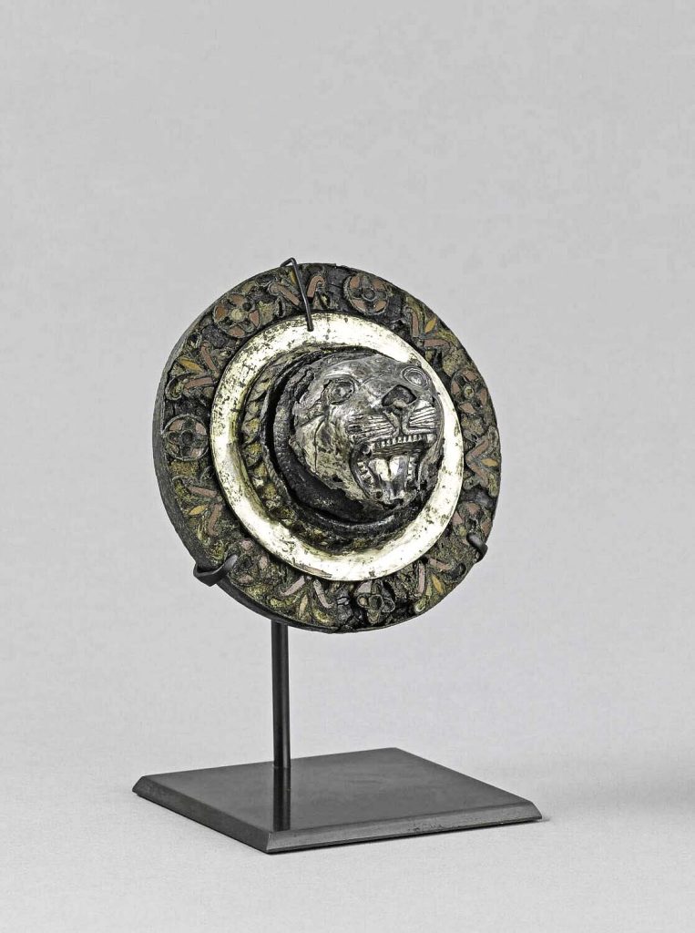 Lion head appliqué decorated with palmettes and rosettes, crafted from bitumen, bronze, and silver.