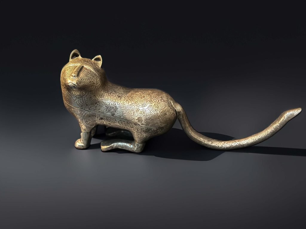 Steel figure of a Persian lion from the Qajar dynasty, intricately engraved with scenes of figures in European dress on a picnic, accompanied by a myriad of birds and beasts, complemented by an enigmatic band of 'square kufic' inscriptions.